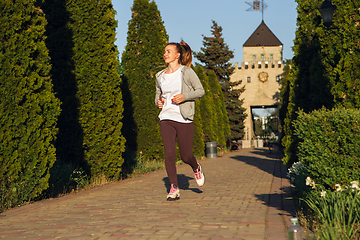 Image showing Young female runner, athlete is jogging in the city street in sunshine. Beautiful caucasian woman training, listening to music