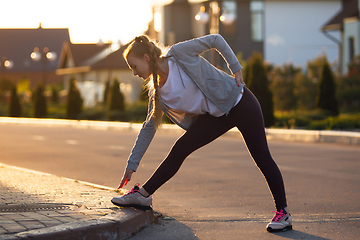 Image showing Young female runner, athlete is stretching before jogging in the city street in sunshine. Beautiful caucasian woman training, listening to music