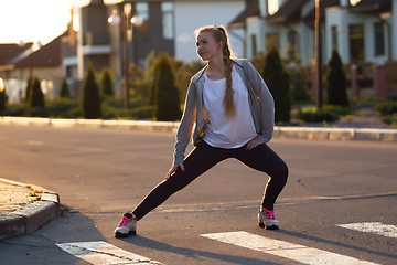 Image showing Young female runner, athlete is stretching before jogging in the city street in sunshine. Beautiful caucasian woman training, listening to music