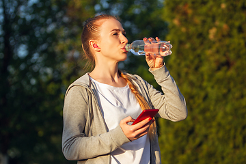 Image showing Young female runner, athlete resting after jogging in the city street in sunshine. Beautiful caucasian woman training, listening to music