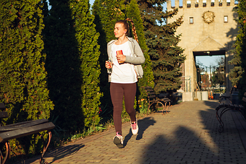 Image showing Young female runner, athlete is jogging in the city street in sunshine. Beautiful caucasian woman training, listening to music