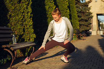 Image showing Young female runner, athlete is stretching before jogging in the city street in sunshine. Beautiful caucasian woman training, listening to music