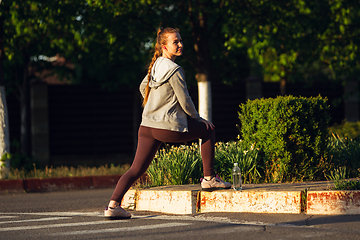 Image showing Young female runner, athlete is stretching before jogging in the city street in sunshine. Beautiful caucasian woman training, listening to music