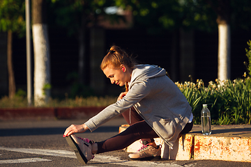 Image showing Young female runner, athlete is stretching before jogging in the city street in sunshine. Beautiful caucasian woman training, listening to music