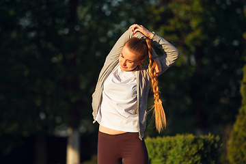 Image showing Young female runner, athlete is stretching before jogging in the city street in sunshine. Beautiful caucasian woman training, listening to music