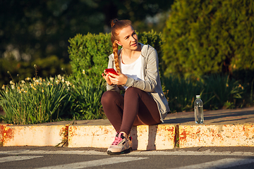 Image showing Young female runner, athlete resting after jogging in the city street in sunshine. Beautiful caucasian woman training, listening to music