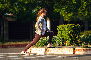 Image showing Young female runner, athlete is stretching before jogging in the city street in sunshine. Beautiful caucasian woman training, listening to music