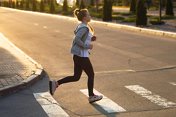 Image showing Young female runner, athlete is jogging in the city street in sunshine. Beautiful caucasian woman training, listening to music