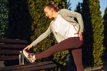 Image showing Young female runner, athlete is stretching before jogging in the city street in sunshine. Beautiful caucasian woman training, listening to music