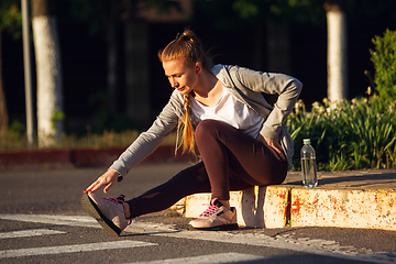 Image showing Young female runner, athlete is stretching before jogging in the city street in sunshine. Beautiful caucasian woman training, listening to music