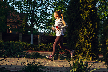 Image showing Young female runner, athlete is jogging in the city street in sunshine. Beautiful caucasian woman training, listening to music