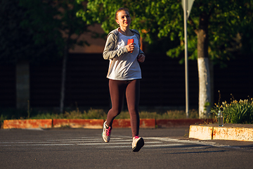 Image showing Young female runner, athlete is jogging in the city street in sunshine. Beautiful caucasian woman training, listening to music