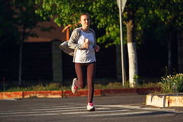 Image showing Young female runner, athlete is jogging in the city street in sunshine. Beautiful caucasian woman training, listening to music