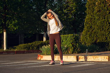 Image showing Young female runner, athlete is stretching before jogging in the city street in sunshine. Beautiful caucasian woman training, listening to music