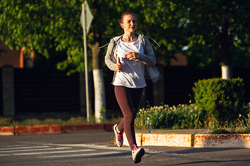 Image showing Young female runner, athlete is jogging in the city street in sunshine. Beautiful caucasian woman training, listening to music