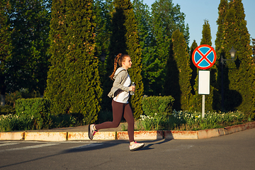 Image showing Young female runner, athlete is jogging in the city street in sunshine. Beautiful caucasian woman training, listening to music