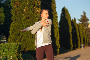 Image showing Young female runner, athlete is stretching before jogging in the city street in sunshine. Beautiful caucasian woman training, listening to music