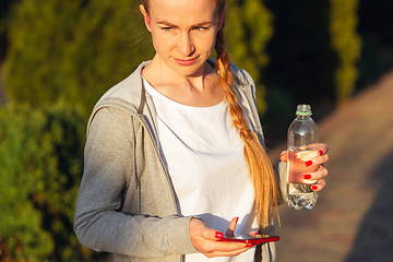 Image showing Young female runner, athlete resting after jogging in the city street in sunshine. Beautiful caucasian woman training, listening to music