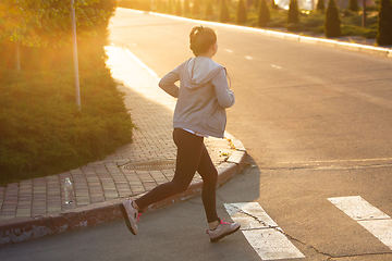 Image showing Young female runner, athlete is jogging in the city street in sunshine. Beautiful caucasian woman training, listening to music