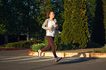 Image showing Young female runner, athlete is jogging in the city street in sunshine. Beautiful caucasian woman training, listening to music