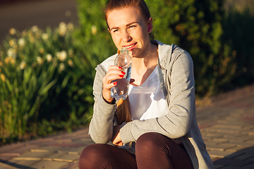 Image showing Young female runner, athlete resting after jogging in the city street in sunshine. Beautiful caucasian woman training, listening to music