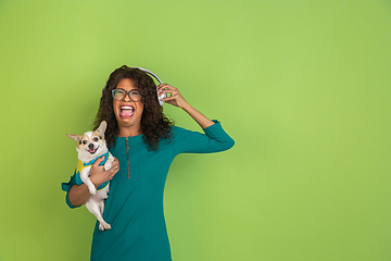 Image showing African-american beautiful young woman\'s portrait with little doggy on green studio background, emotional and expressive. Copyspace for ad.