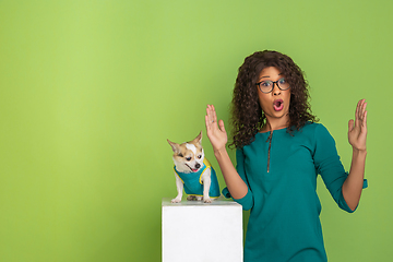 Image showing African-american beautiful young woman\'s portrait with little doggy on green studio background, emotional and expressive. Copyspace for ad.