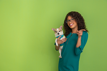 Image showing African-american beautiful young woman\'s portrait with little doggy on green studio background, emotional and expressive. Copyspace for ad.