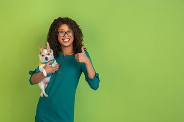 Image showing African-american beautiful young woman\'s portrait with little doggy on green studio background, emotional and expressive. Copyspace for ad.