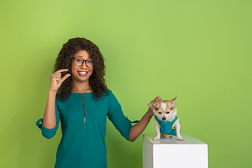 Image showing African-american beautiful young woman\'s portrait with little doggy on green studio background, emotional and expressive. Copyspace for ad.