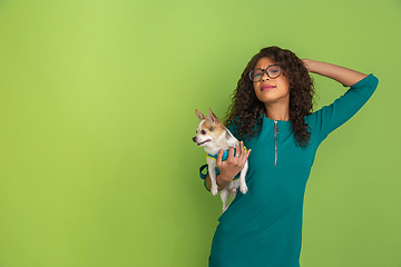 Image showing African-american beautiful young woman\'s portrait with little doggy on green studio background, emotional and expressive. Copyspace for ad.