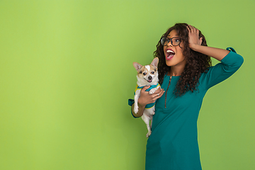 Image showing African-american beautiful young woman\'s portrait with little doggy on green studio background, emotional and expressive. Copyspace for ad.