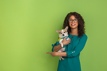 Image showing African-american beautiful young woman\'s portrait with little doggy on green studio background, emotional and expressive. Copyspace for ad.