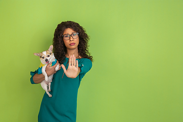 Image showing African-american beautiful young woman\'s portrait with little doggy on green studio background, emotional and expressive. Copyspace for ad.