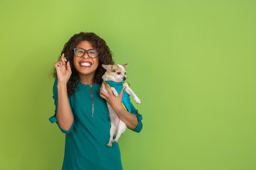 Image showing African-american beautiful young woman\'s portrait with little doggy on green studio background, emotional and expressive. Copyspace for ad.