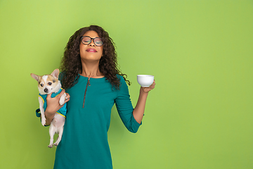 Image showing African-american beautiful young woman\'s portrait with little doggy on green studio background, emotional and expressive. Copyspace for ad.