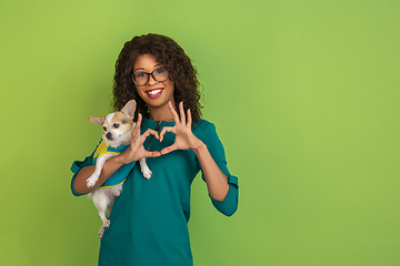 Image showing African-american beautiful young woman\'s portrait with little doggy on green studio background, emotional and expressive. Copyspace for ad.