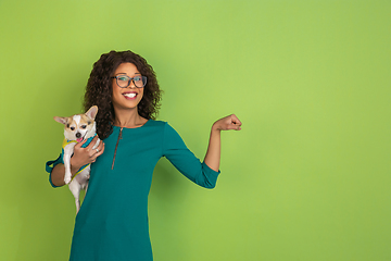 Image showing African-american beautiful young woman\'s portrait with little doggy on green studio background, emotional and expressive. Copyspace for ad.