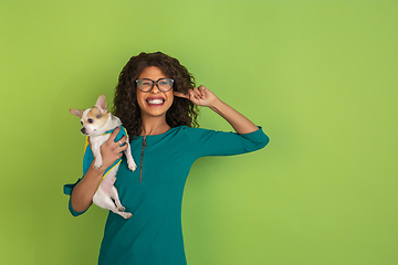 Image showing African-american beautiful young woman\'s portrait with little doggy on green studio background, emotional and expressive. Copyspace for ad.