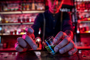 Image showing Barman finishes preparation of alcoholic cocktail, sets fire to drink in multicolored neon light, focus on glass