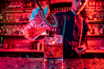 Image showing Barman finishes preparation of alcoholic cocktail, pouring drink in multicolored neon light, focus on glass