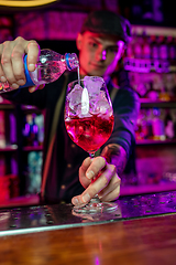 Image showing Close up of barman finishes preparation of alcoholic cocktail, pouring drink in multicolored neon light, focus on glass