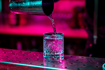 Image showing Close up of barman finishes preparation of alcoholic cocktail, pouring drink in multicolored neon light