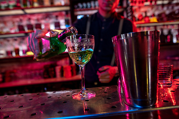 Image showing Barman finishes preparation of alcoholic cocktail, pouring drink in multicolored neon light, focus on glass