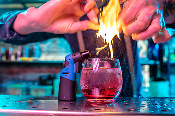 Image showing Close up of alcoholic cocktail, beverage, drink in multicolored neon light with fire flames setting on orange\'s spraying juice
