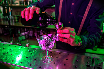 Image showing Close up of barman finishes preparation of alcoholic cocktail, pouring drink with shot in multicolored neon light, focus on glass
