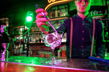 Image showing Close up of barman finishes preparation of alcoholic cocktail, pouring drink in multicolored neon light, focus on glass