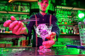 Image showing Close up of barman finishes preparation of alcoholic cocktail, pouring drink in multicolored neon light, focus on glass