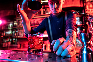 Image showing Close up of barman finishes preparation of alcoholic cocktail, pouring drink in multicolored neon light, focus on glass