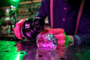 Image showing Close up of barman finishes preparation of alcoholic cocktail, pouring drink in multicolored neon light, focus on glass